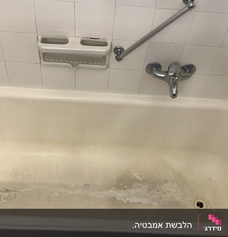 אמבטיה לבנה עם כתמים, ברז ומחזיק סבון על קיר אריחים לבנים.